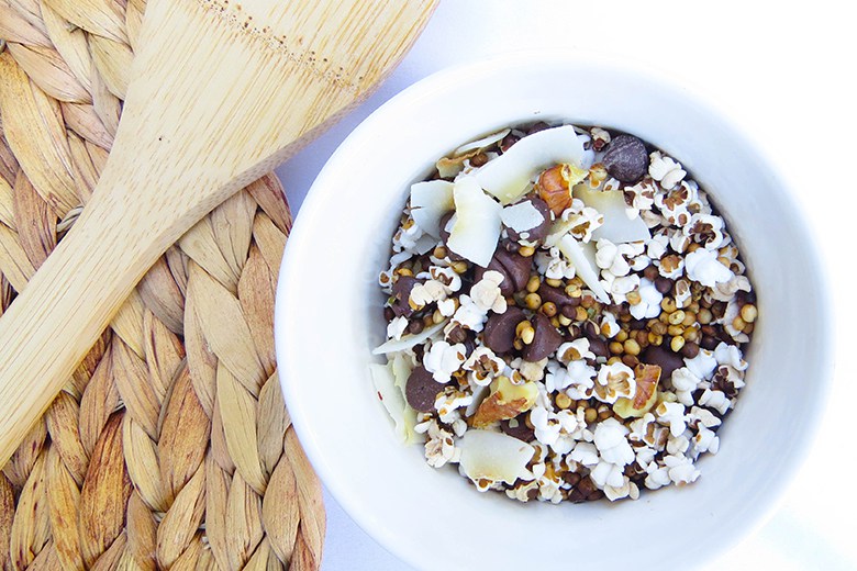 Popped Sorghum Trail Mix in white bowl next to a woven placemat and wooden spoon