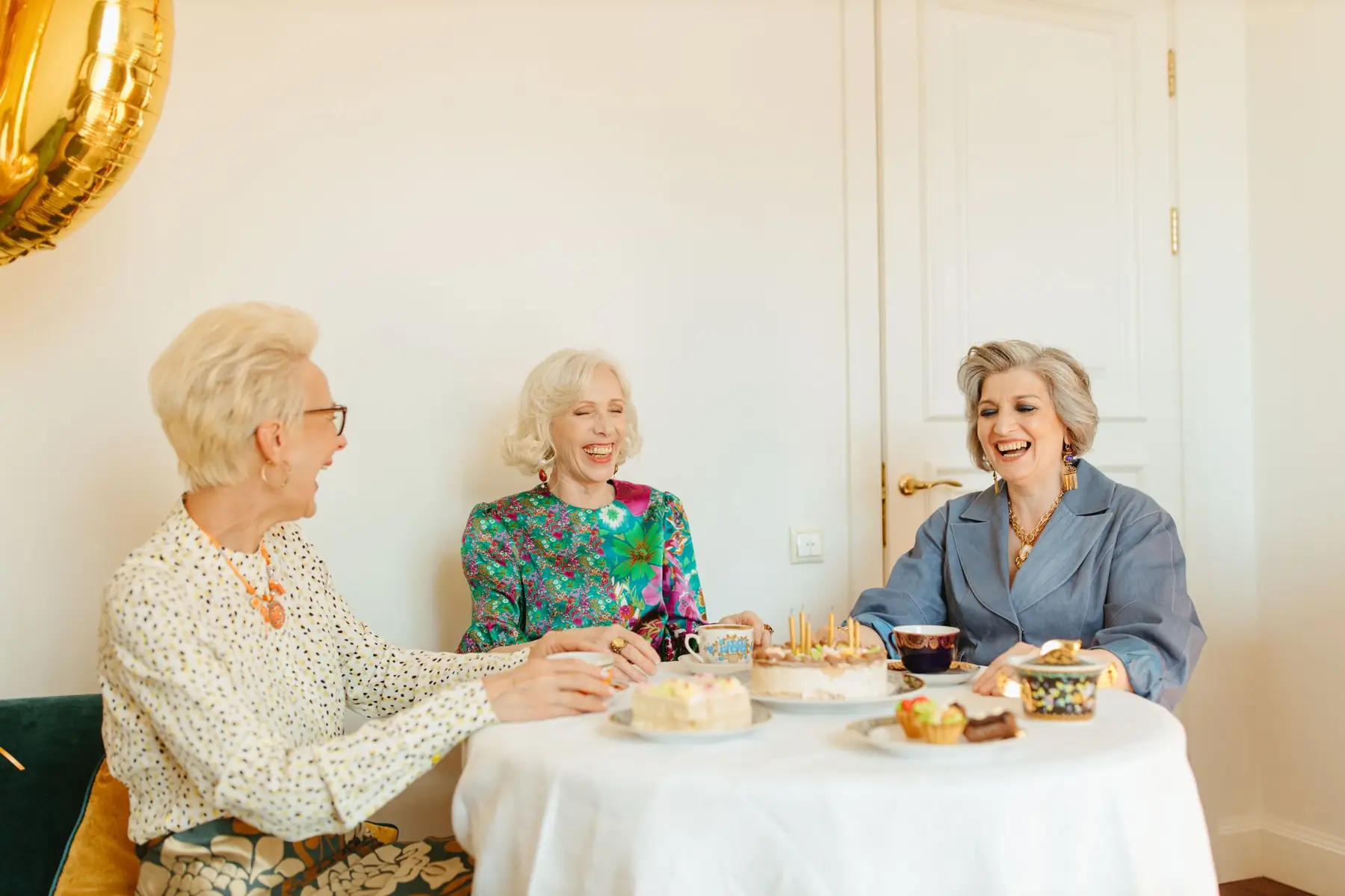 Three senior women laughing and enjoying a tea party with cakes indoors