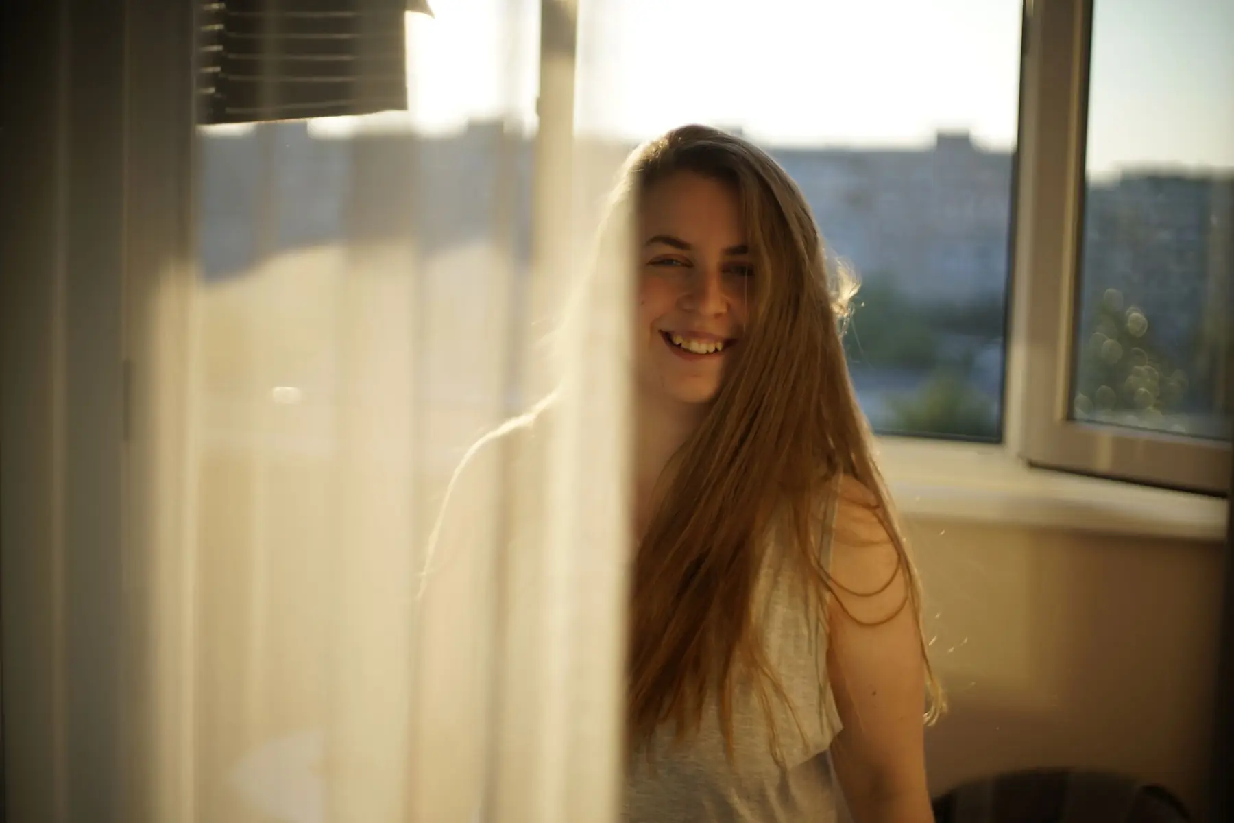A young woman smiles warmly by a window as sunlight streams through a curtain