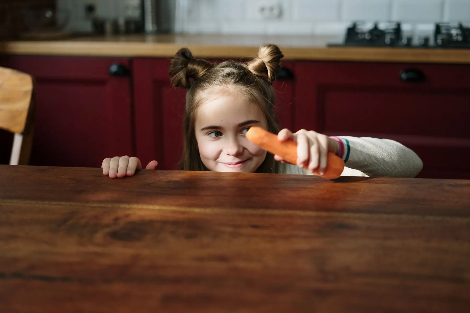 Playful child with a carrot peeking over a wooden table in a cozy kitchen