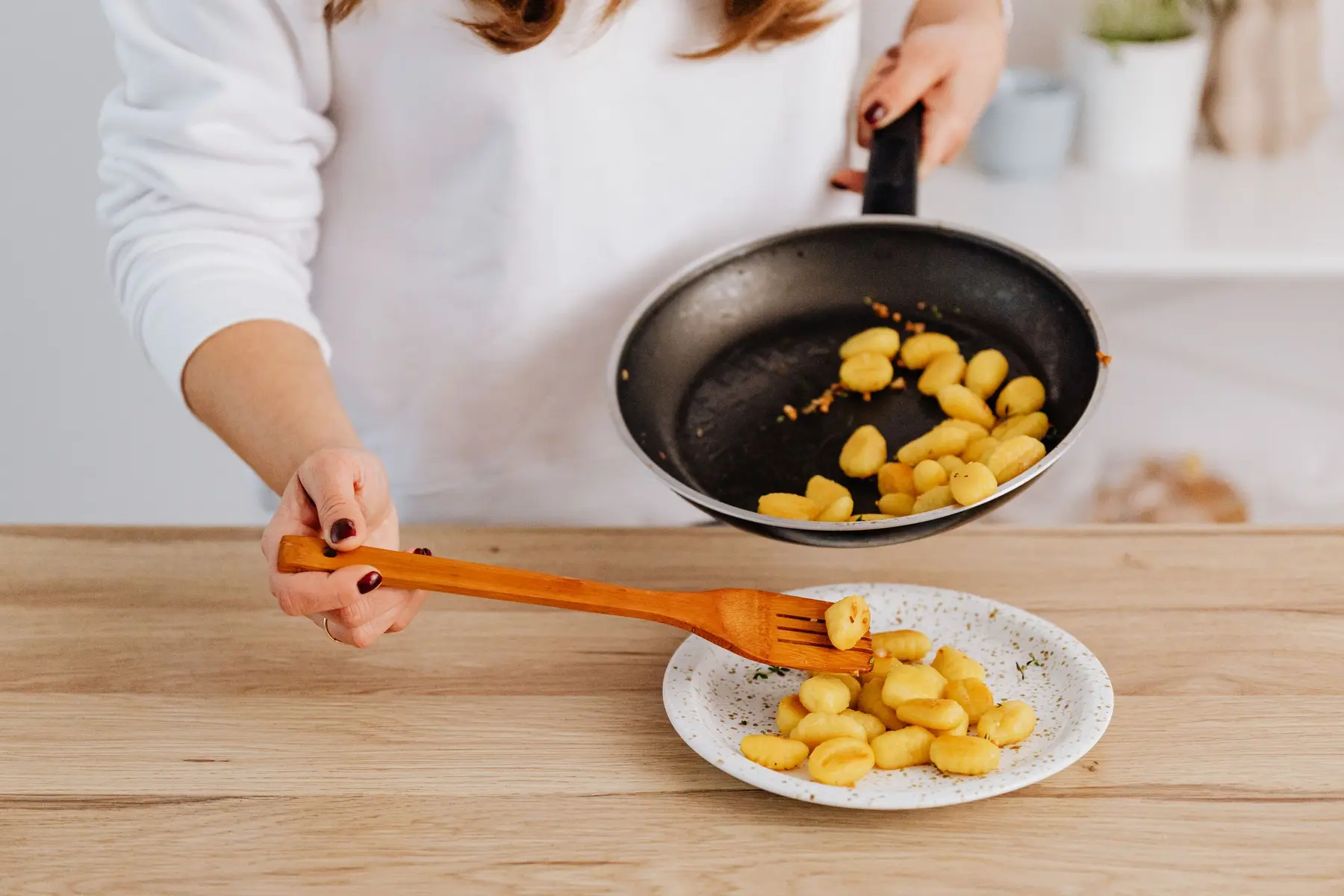 Hands plating golden gnocchi from a pan onto a plate on wood
