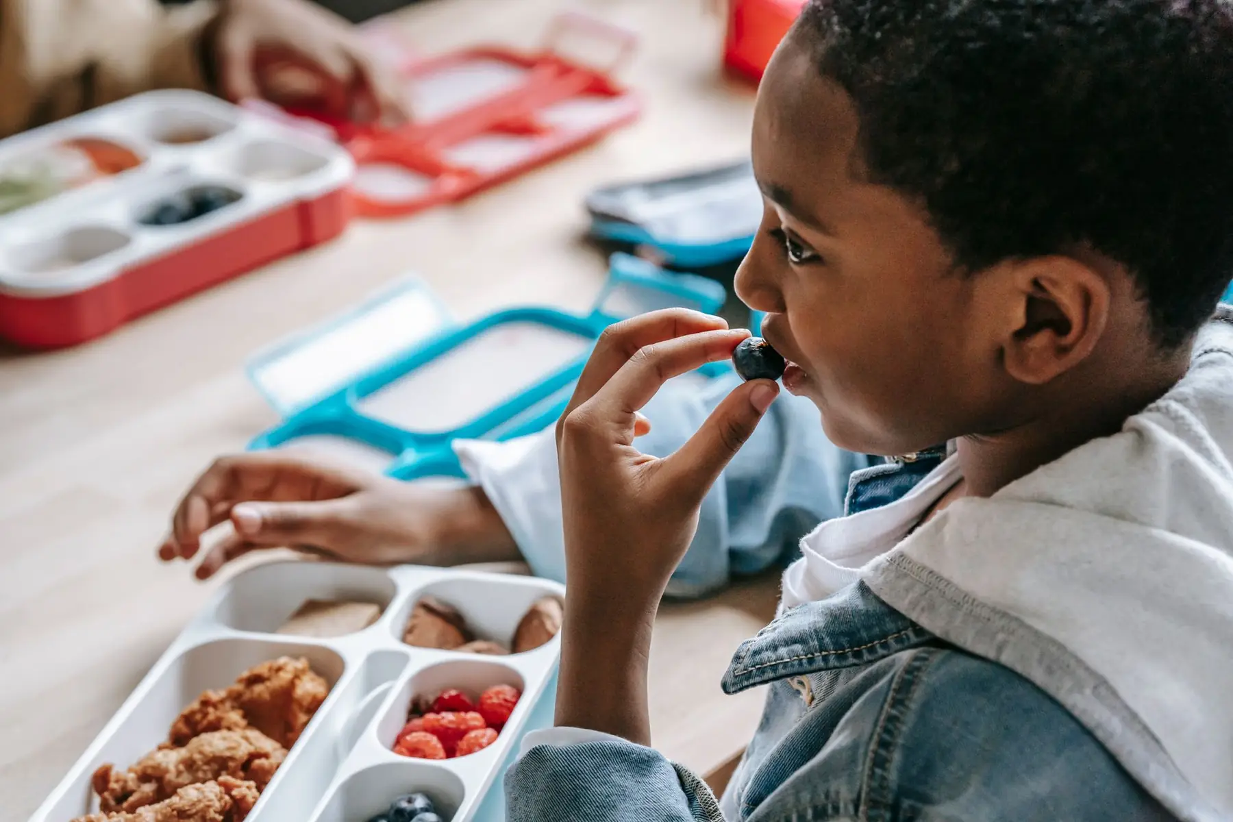 Crop African American child in casual clothes sitting at table with plastic lunch box