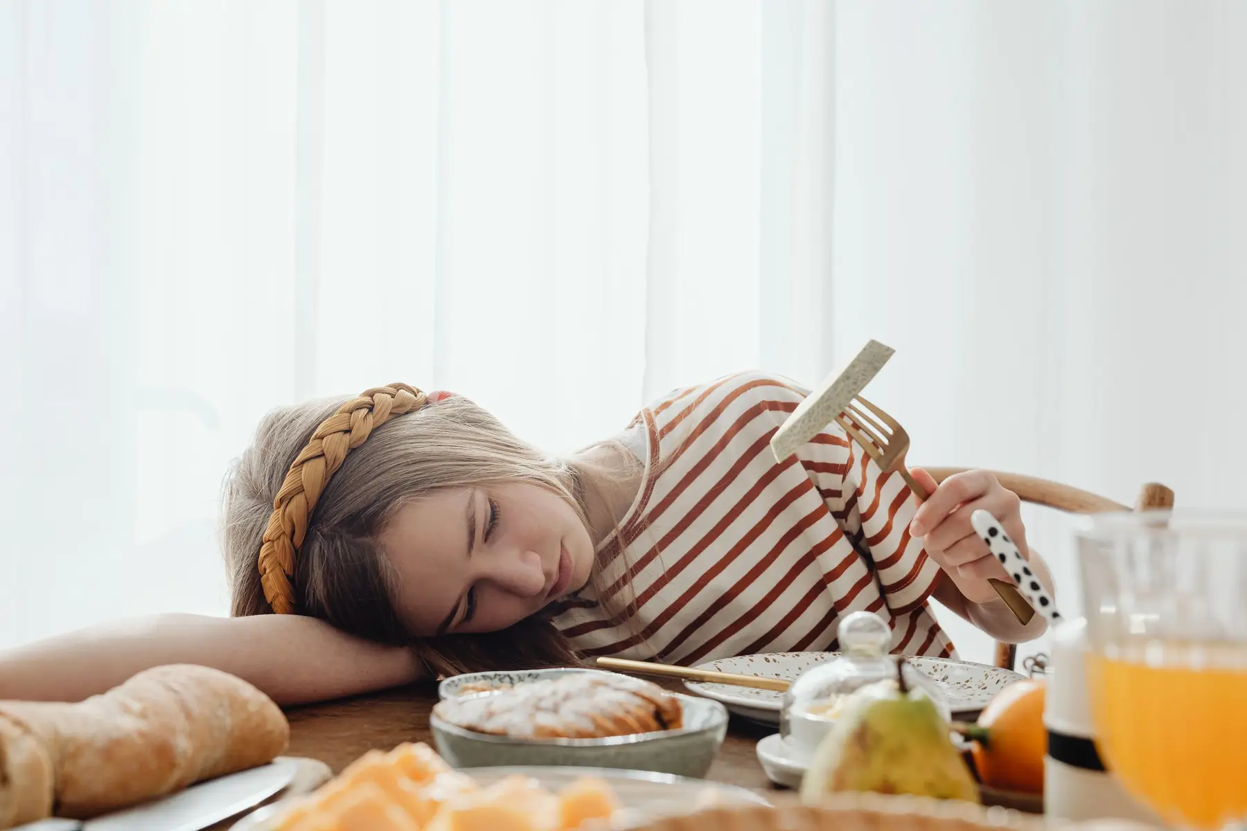 A tired woman rests her head on a sunny breakfast table