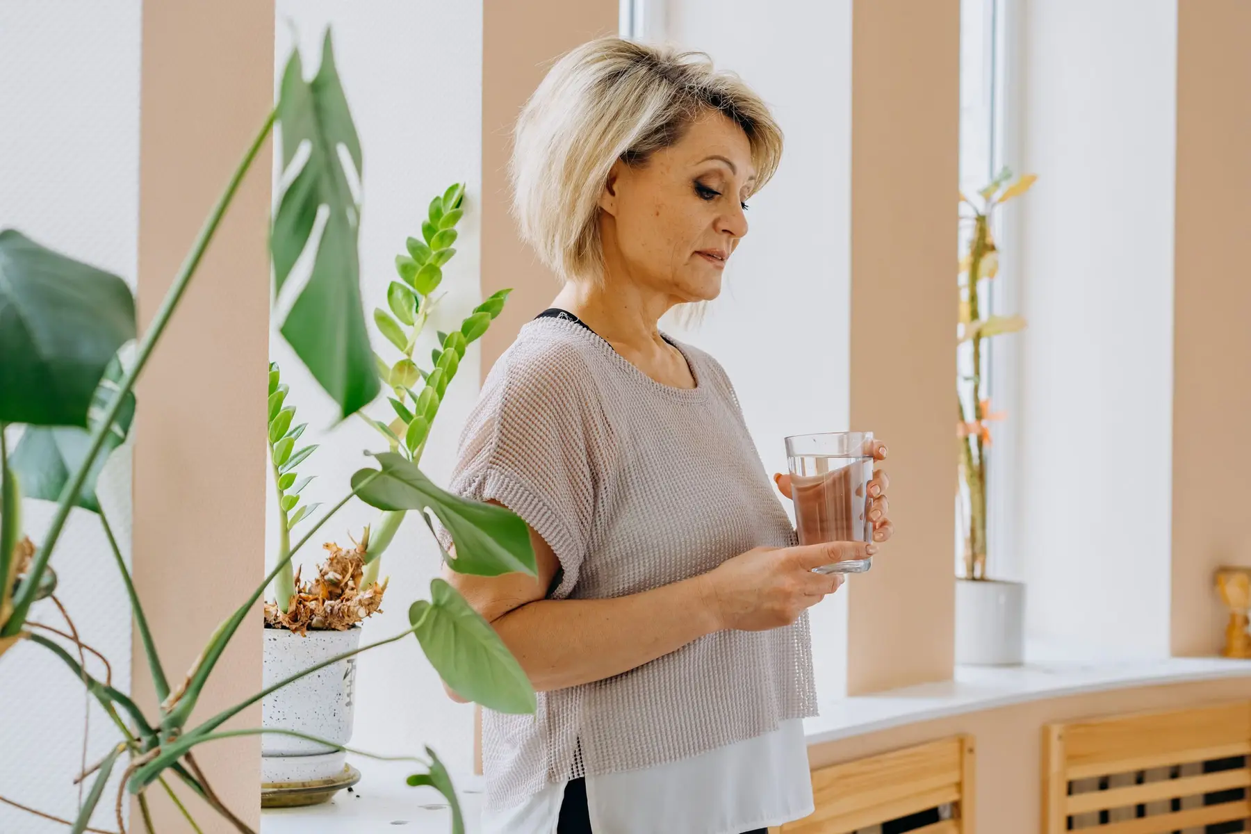 A mature woman holds a glass of water among green plants at home