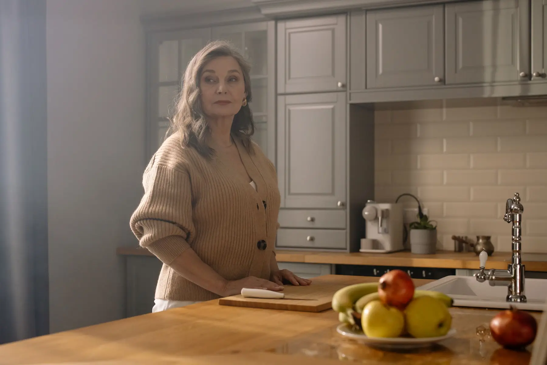 A woman stands at a wooden kitchen counter beside a bowl of fruit