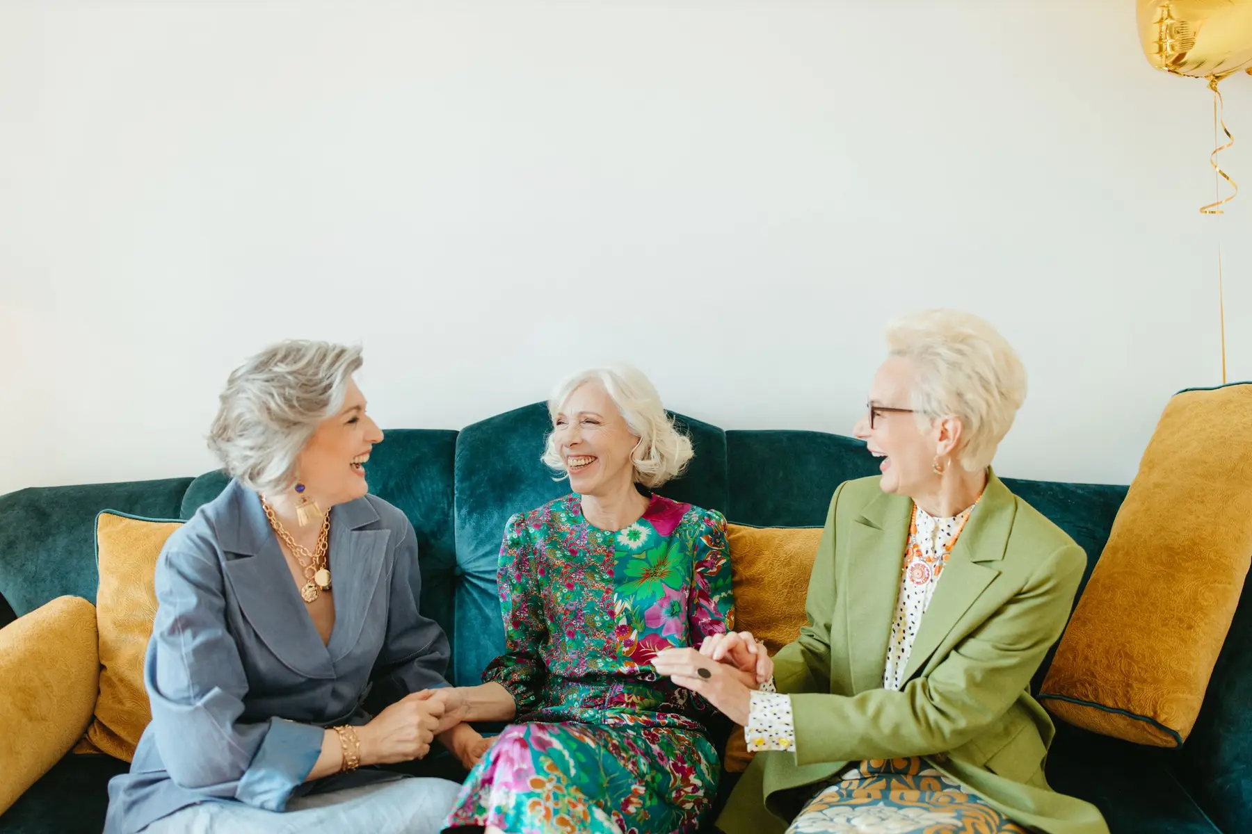 Three older women laugh together on a cozy sofa
