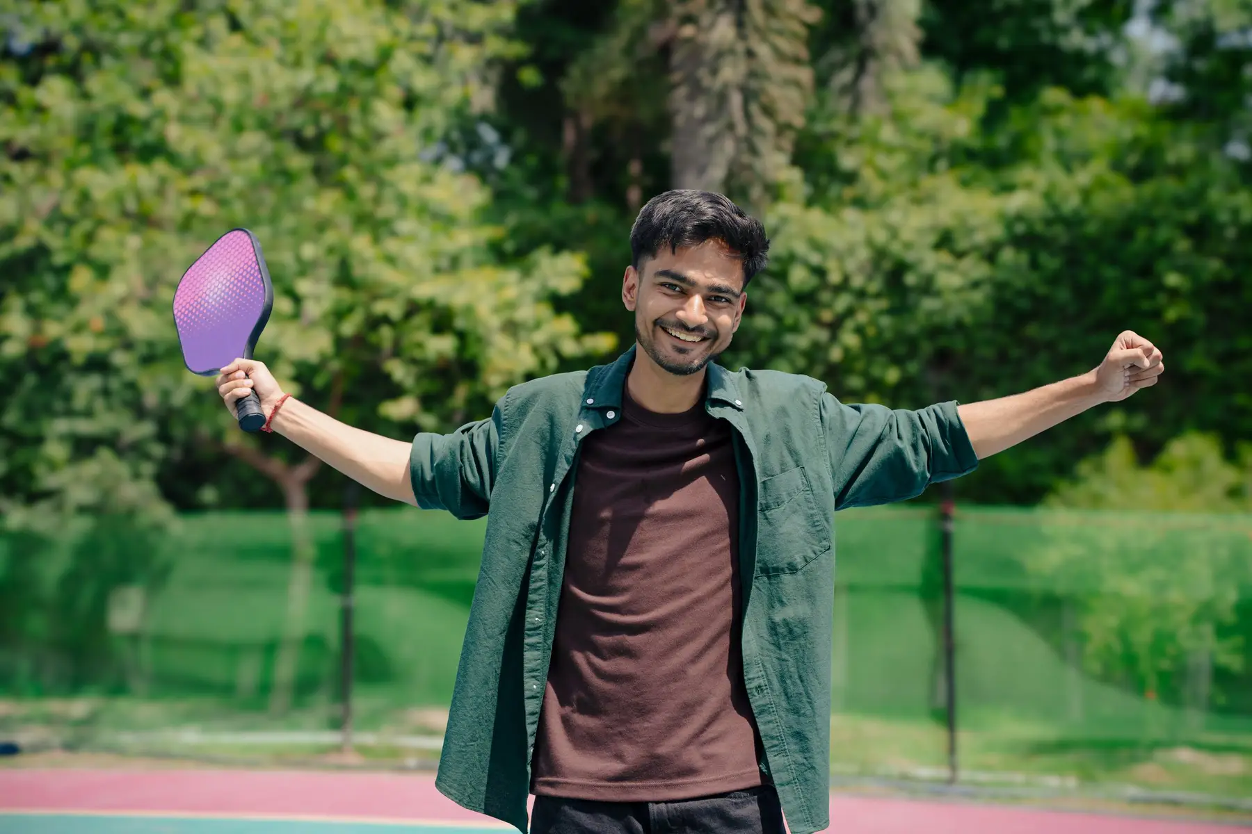 A cheerful man raises his pickleball paddle with arms wide outdoors