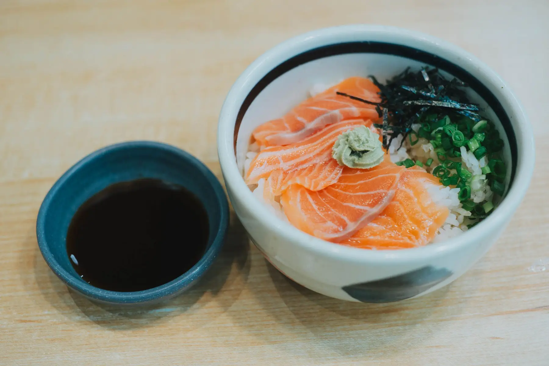 A salmon rice bowl beside a small dish of soy sauce on light wood