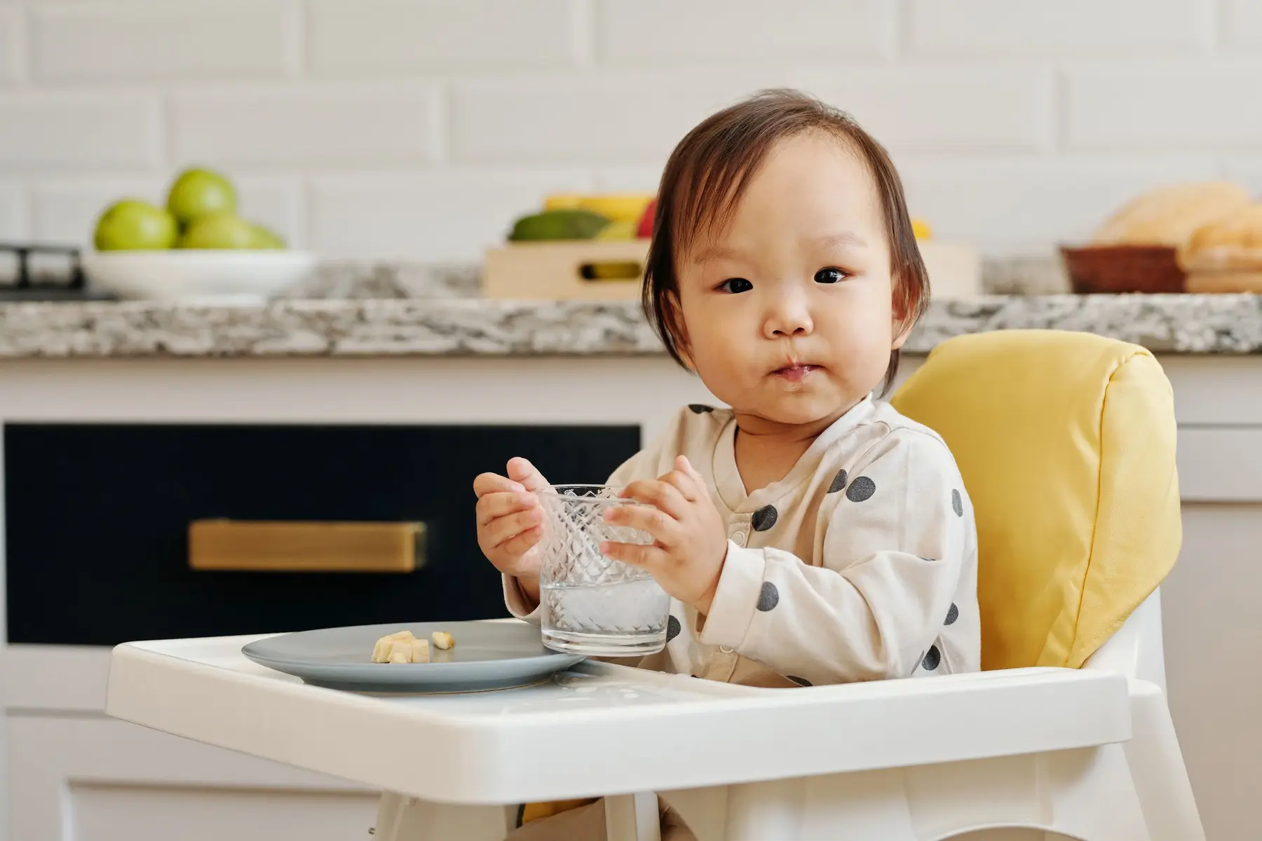 A toddler sips water from a small glass in a sunny kitchen