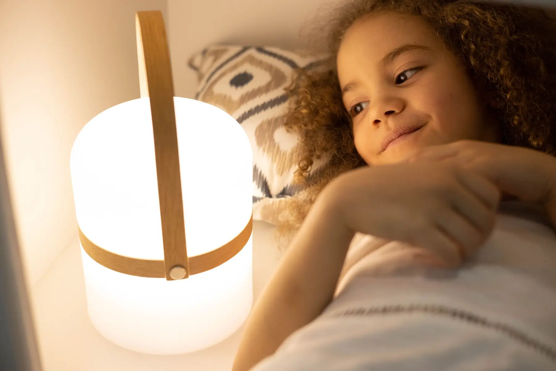 A smiling child lies in bed beside a softly glowing lamp