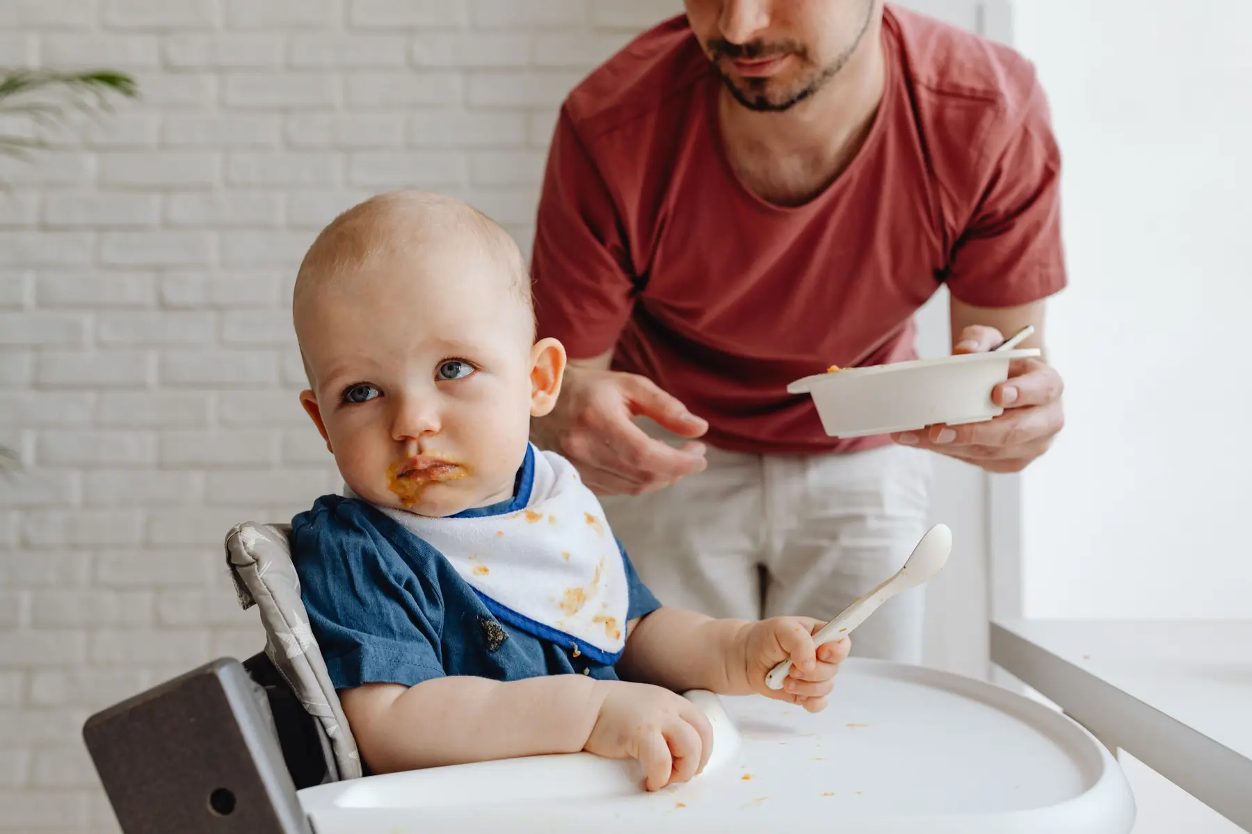 A toddler in a high chair looks unsure as a parent offers a bowl