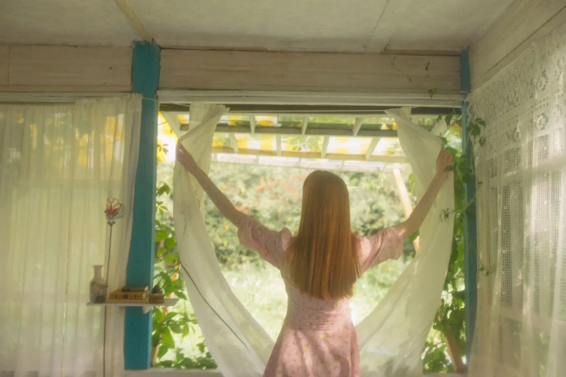 A woman opens sheer curtains to let morning light into the room
