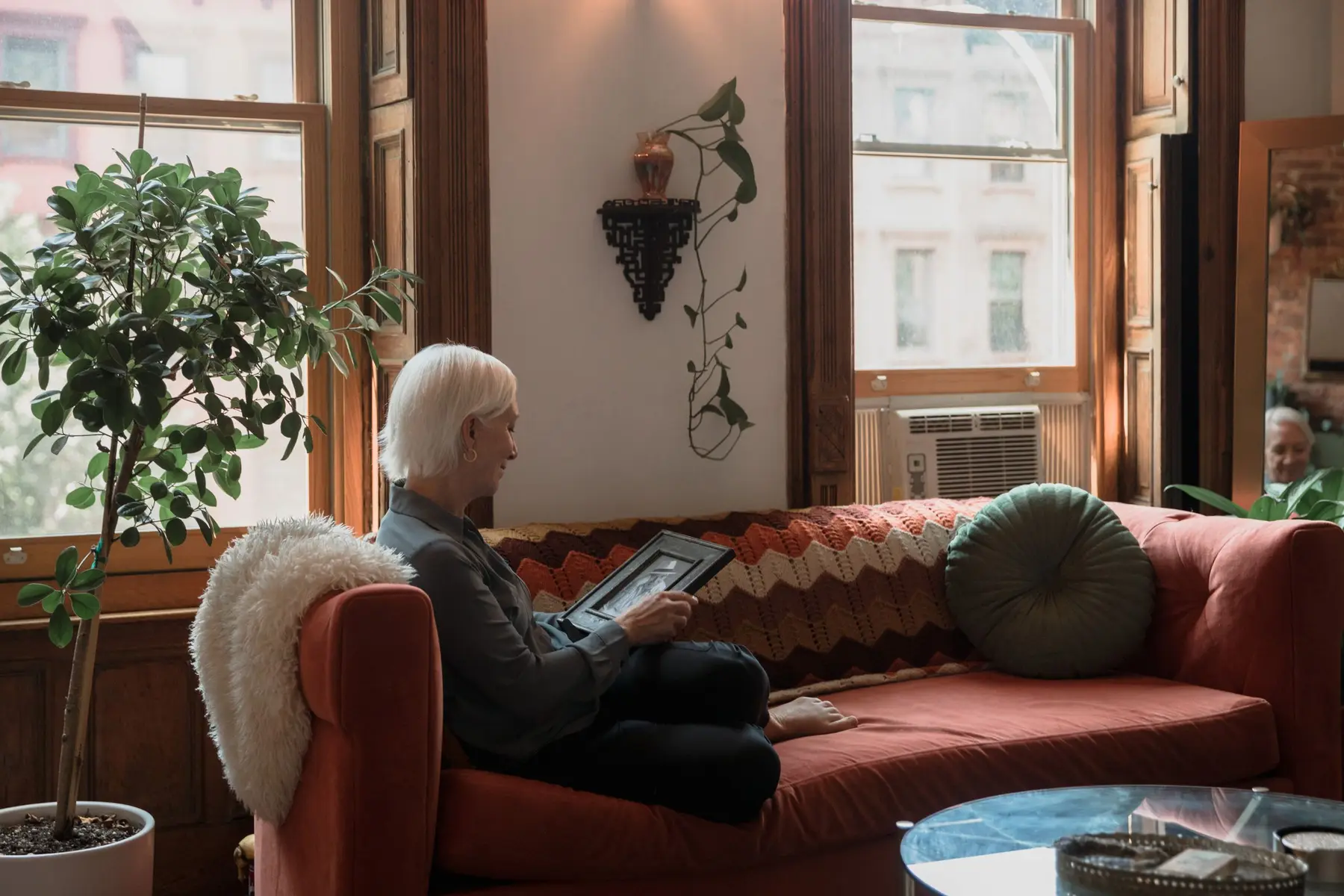 An older woman sits on a red sofa in a sunlit room