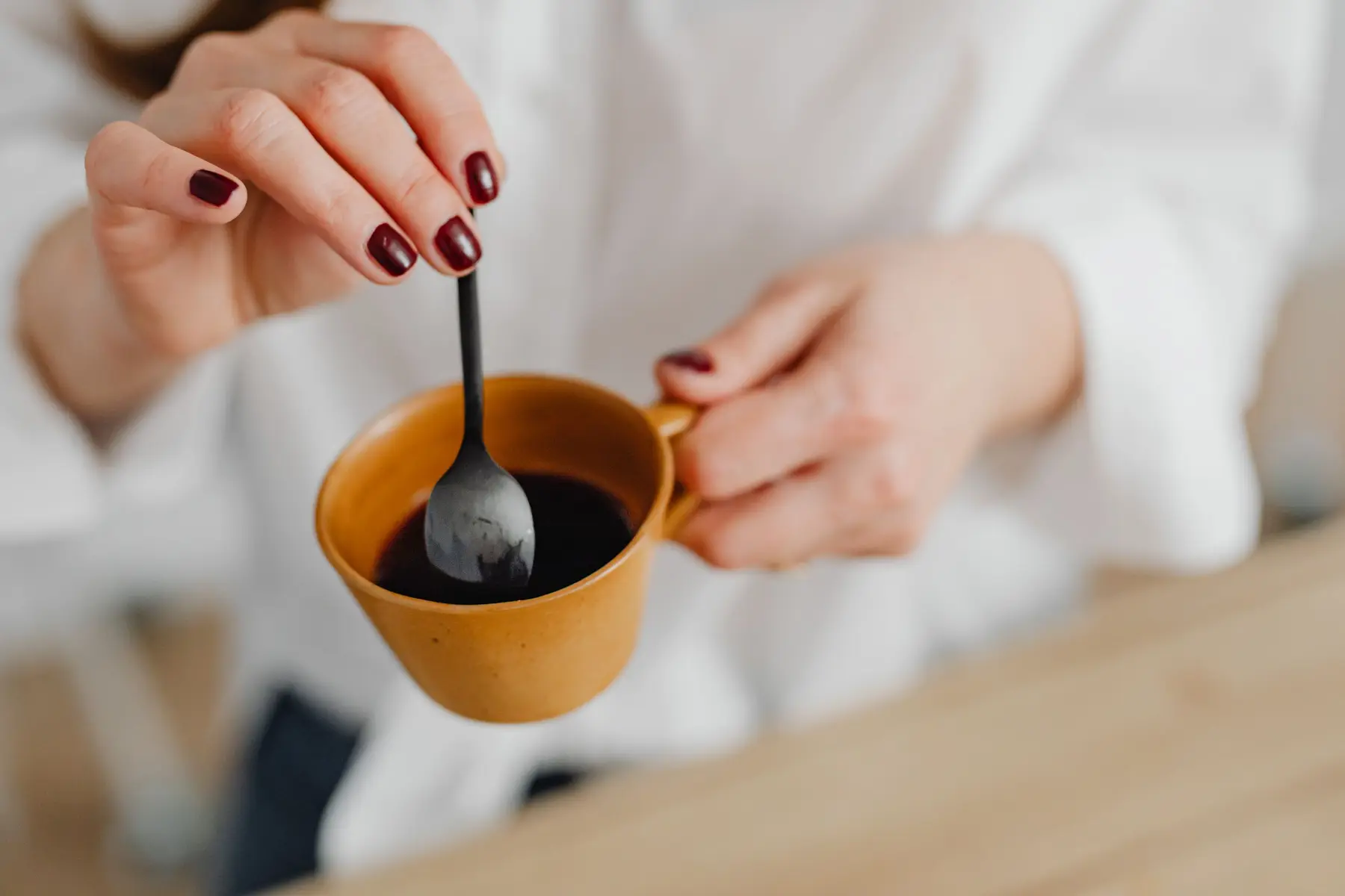 A woman stirs a warm drink in an orange mug at a wooden table