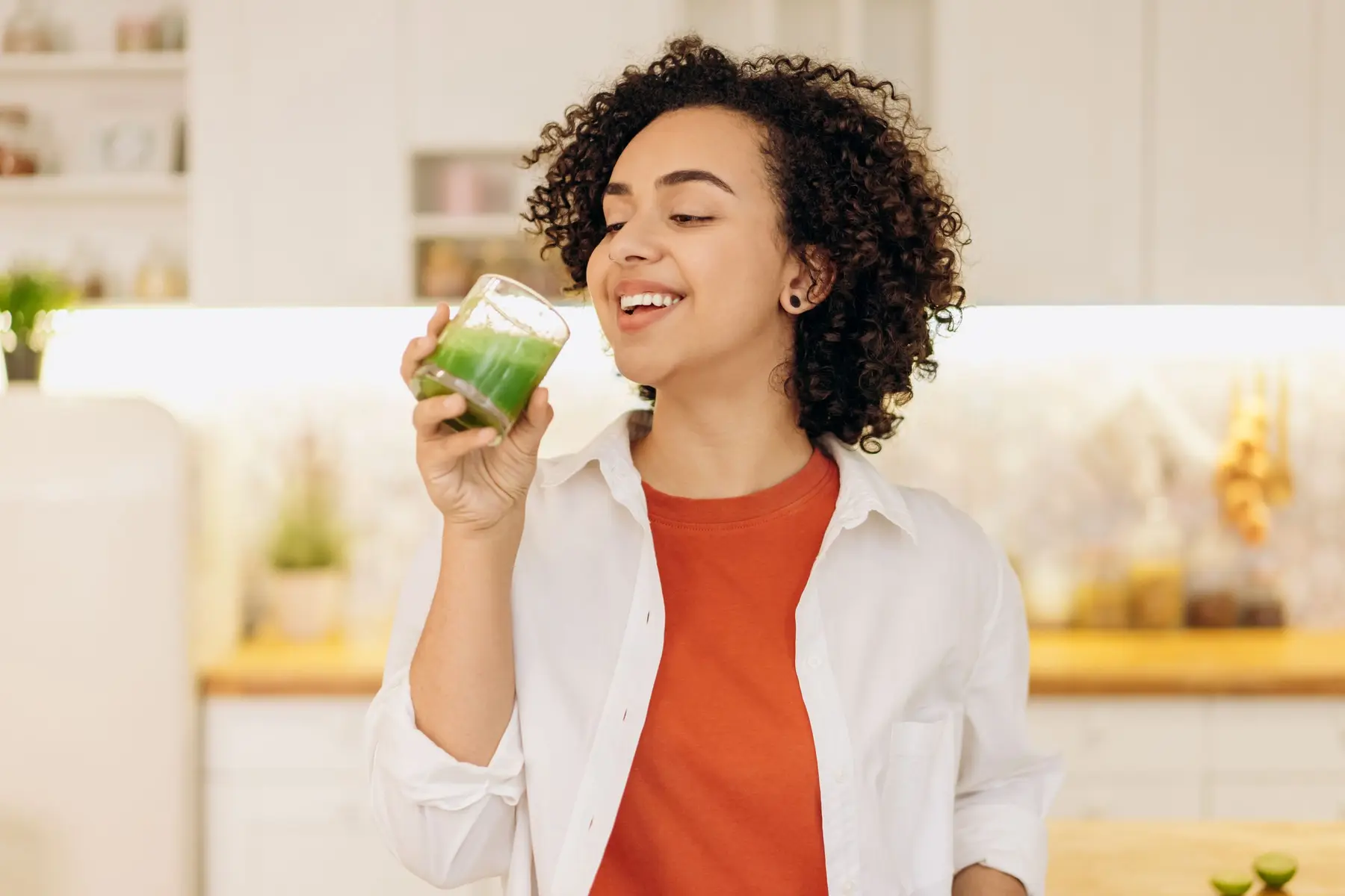 A smiling woman sips a green smoothie in a bright kitchen