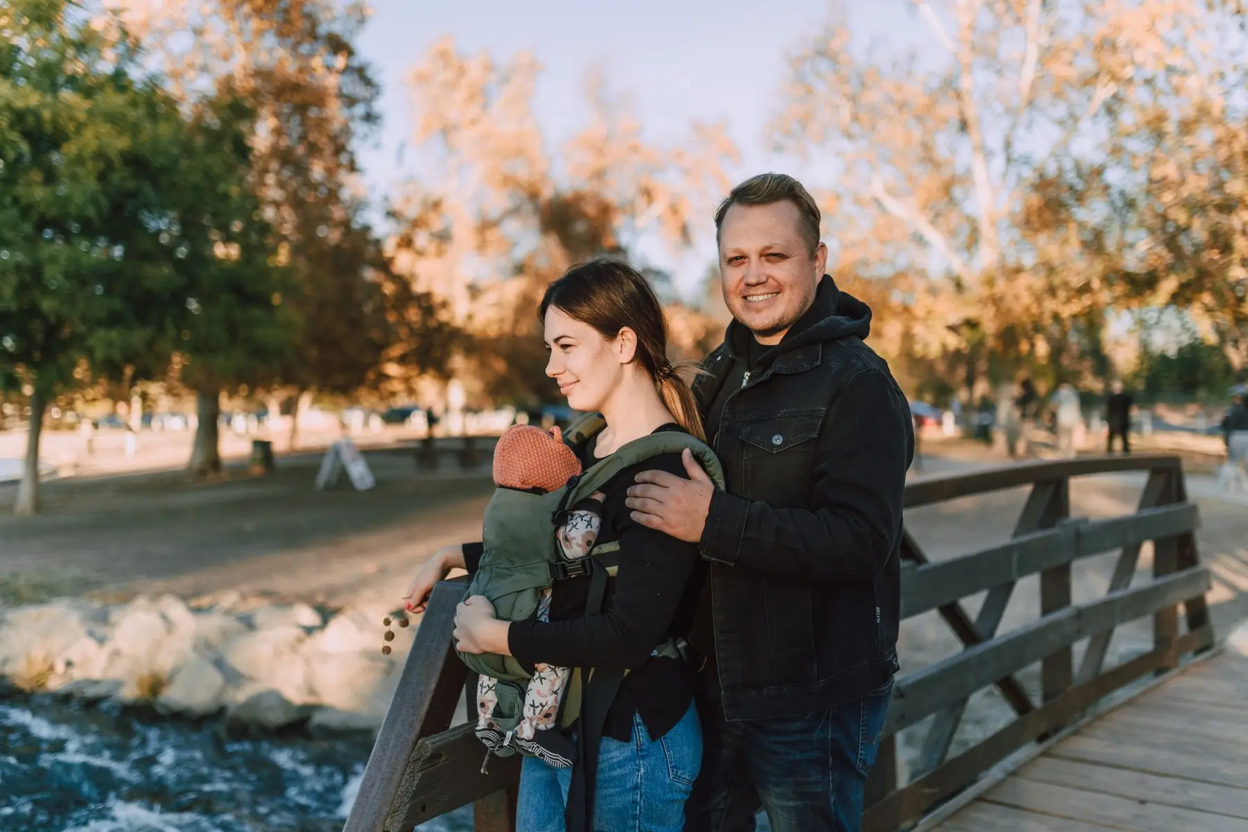 A joyful couple with a baby in a carrier at a scenic park bridge