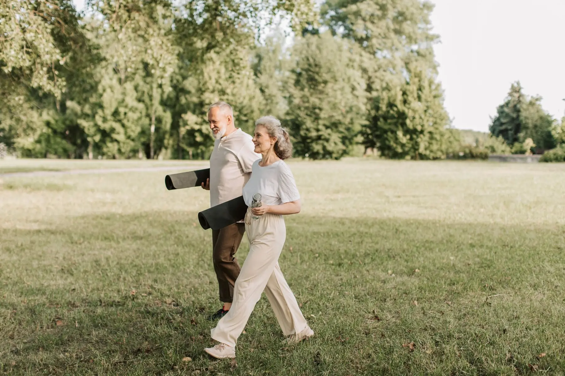 Elderly couple walking on grass carrying yoga mats, enjoying a sunny day outdoors