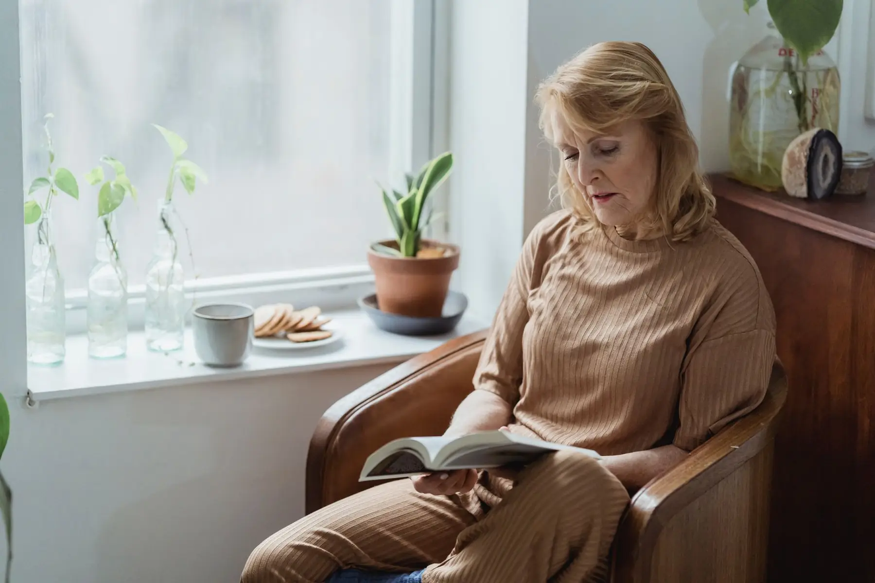 Senior female reading book in armchair against mug of hot beverage and tasty biscuits