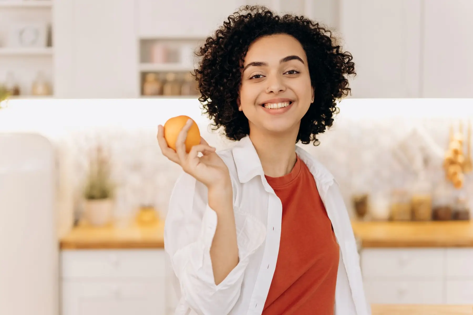 A joyful woman with curly hair holding an orange in her kitchen, promoting a